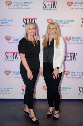 Two women posing and smiling at an event, standing in front of a step-and-repeat banner with logos for Nicklaus Children’s Health System and SFBW (South Florida Business & Wealth). Both wear professional black and white attire. South Florida Business & Wealth