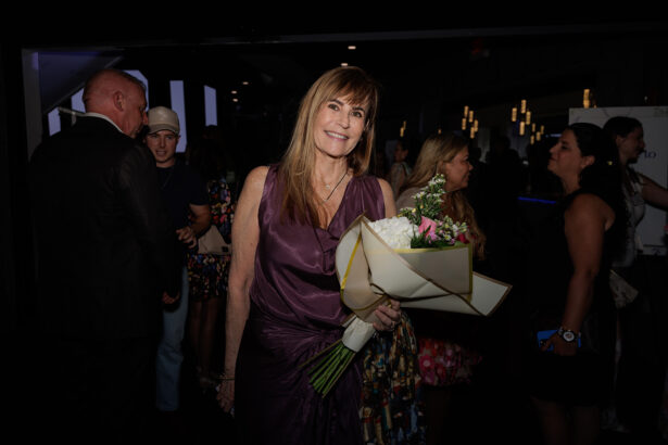 A woman in a purple dress holds a bouquet of flowers and smiles at the camera. She is standing at an indoor event with several people in the background. The lighting is dim, emphasizing her presence. South Florida Business & Wealth