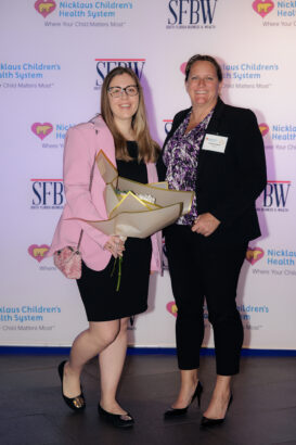 Two women dressed in business attire smile at the camera. One holds a bouquet of flowers. They stand in front of a backdrop with logos for Nicklaus Children's Health System and SFBW. South Florida Business & Wealth