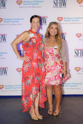 Two women posing and smiling in front of a step-and-repeat banner with logos for Nicklaus Children’s Health System and South Florida Business & Wealth. Both are wearing colorful dresses and name badges. South Florida Business & Wealth