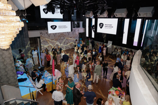 A large group of people socializes at an indoor event with tables, decorations, and a "Recovery Unplugged" logo displayed on multiple screens above the crowd. The setting is modern with bright lighting and a chandelier. South Florida Business & Wealth