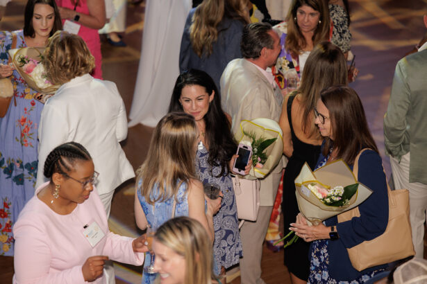 A group of people, mostly women, are gathered indoors, conversing and holding bouquets of flowers. Several wear name tags and dresses, suggesting a formal or celebratory event. South Florida Business & Wealth