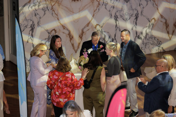 A group of people stand around a small round table at an indoor event, talking and smiling. The setting features a marble-patterned wall and patterned lighting on the floor. Some hold drinks and flowers. South Florida Business & Wealth