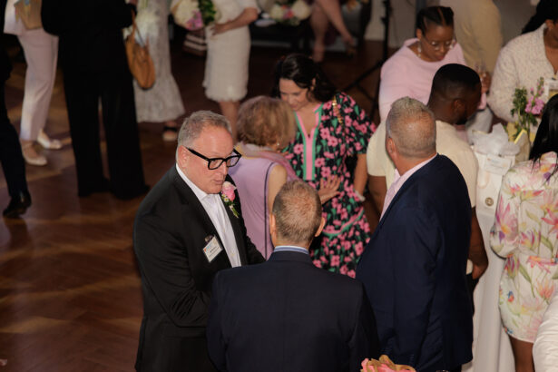 A group of people, dressed in formal and semi-formal attire, are mingling and conversing at an indoor event with wooden floors. Some hold drinks and flowers; the atmosphere appears social and lively. South Florida Business & Wealth
