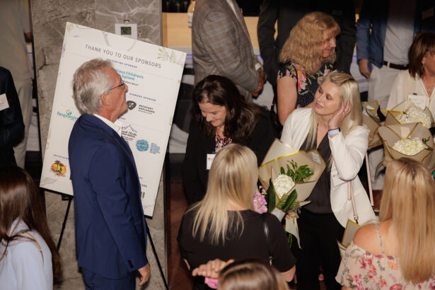A group of people in formal attire gather at an event. Some hold bouquets and chat, while a man in a blue suit stands near a sponsor board. The atmosphere is lively and social. South Florida Business & Wealth