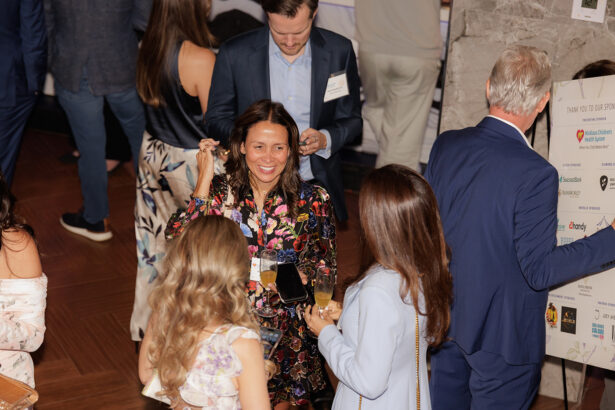A group of people in formal attire socialize at an indoor event, holding drinks and smiling. One woman in a floral dress is talking with two women, while a man in a blue suit looks at a board displaying sponsor logos. South Florida Business & Wealth