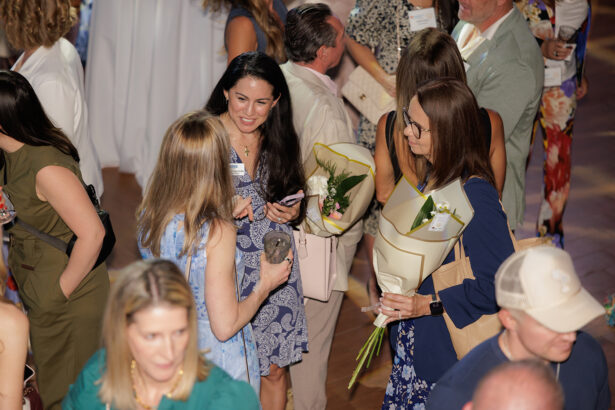 A group of people, mostly women, are gathered indoors at a social event. Some are talking and holding drinks, while one woman holds flowers and a folder. The atmosphere is lively and casual. South Florida Business & Wealth
