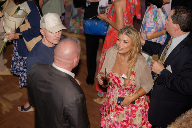 A group of people at an indoor social event talk and mingle. A woman in a pink floral dress holds a drink and smiles, standing with three men. Others are visible in the background, dressed in colorful attire. South Florida Business & Wealth