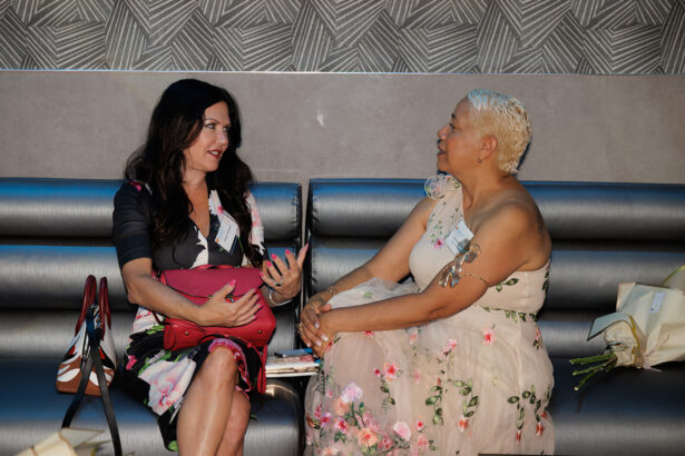 Two women sit on a silver bench, engaged in conversation. One has long dark hair and a floral dress, the other has short blonde hair and a light floral dress. Bouquets of flowers rest beside them. South Florida Business & Wealth