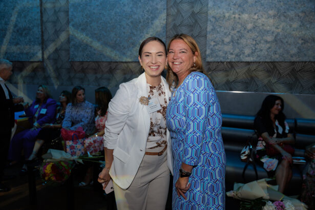 Two women smiling and posing together at an indoor event, with a group of people seated in the background. The setting appears formal, and there are bouquets of flowers on the bench beside the seated guests. South Florida Business & Wealth