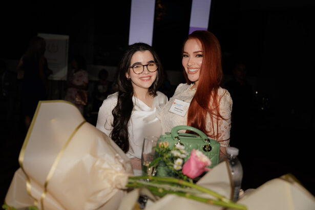 Two women dressed in white smiling at an event table decorated with large flower bouquets, a green purse, a water bottle, and glassware, with a dark background and blurred people behind them. South Florida Business & Wealth