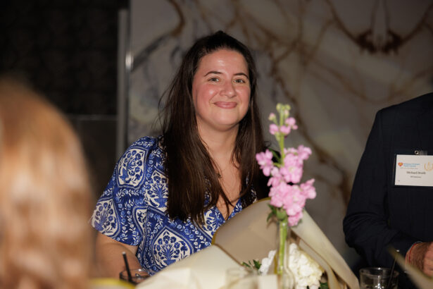 A woman with long dark hair, wearing a blue patterned dress, smiles while holding a bouquet of flowers at an indoor event. She is standing near a table with drinks and a pink flower centerpiece. South Florida Business & Wealth