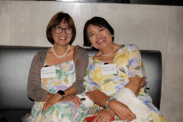 Two smiling women wearing glasses and floral dresses sit closely together on a bench, each with a name tag and holding handbags, posing for a photo in a well-lit indoor setting. South Florida Business & Wealth