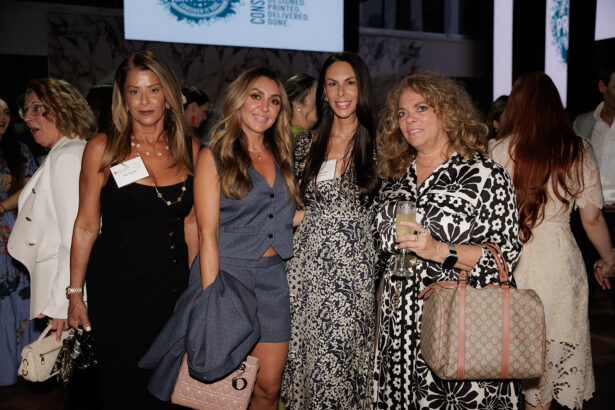 Four women in stylish outfits pose together at an indoor event. One holds a drink and another holds a jacket. Other attendees and large screens are visible in the background. South Florida Business & Wealth