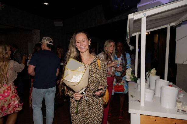 A woman in a patterned dress smiles and holds a bouquet of flowers at an indoor event, with other people socializing in the background. Nearby is a white booth or stand displaying several white containers. South Florida Business & Wealth
