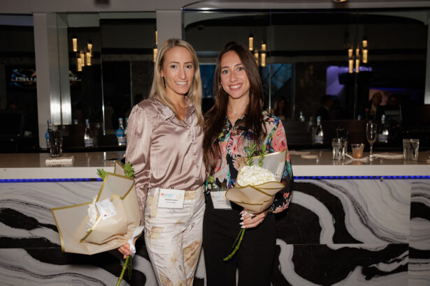 Two women stand side by side indoors, smiling at the camera and holding bouquets of white flowers. They wear business casual attire and name badges, with a modern bar and hanging lights in the background. South Florida Business & Wealth