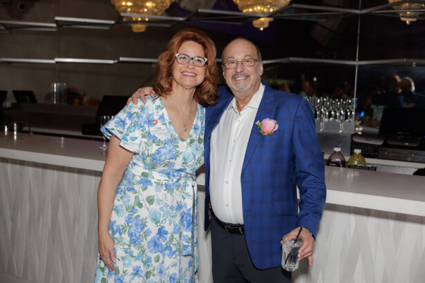 A smiling woman in a white and blue floral dress stands next to a smiling man in a blue plaid jacket holding a drink, both posing in front of a modern white bar with mirrors and chandeliers in the background. South Florida Business & Wealth