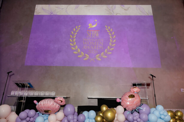 A stage decorated with pastel balloon arrangements and large artificial flowers, with two podiums. On the wall, a purple projection reads, “SFBW Prestigious Women Awards 2026” surrounded by a gold laurel wreath. South Florida Business & Wealth