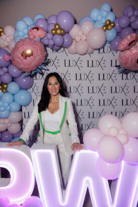 A woman in a white suit with green accents stands in front of a Luxe backdrop, surrounded by pastel balloons and large illuminated letters. The setting appears festive, with pink, purple, and blue decorations. South Florida Business & Wealth