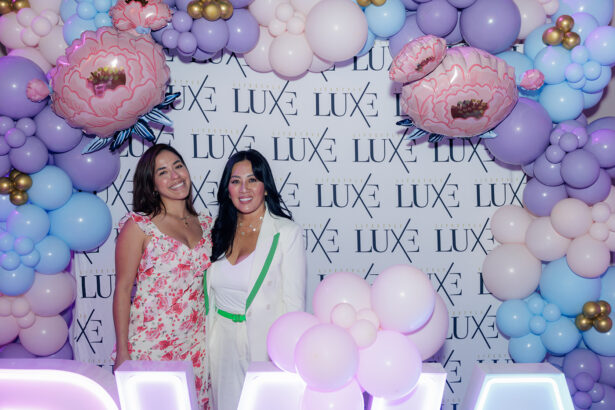 Two women in floral dresses smile and pose together in front of a step-and-repeat backdrop reading “LUXE,” surrounded by pastel and gold balloon decorations and large pink flower balloons. South Florida Business & Wealth