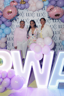 Three women stand smiling in front of a decorative balloon arch and a wall with “LUXE” repeated on it. Large illuminated letters and pastel-colored balloons create a festive atmosphere. South Florida Business & Wealth