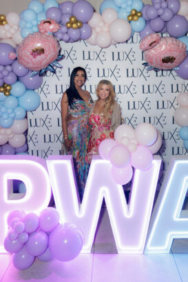 Two women in floral dresses stand smiling in front of a “LUXE” backdrop, surrounded by pastel balloon decorations and illuminated “PWA” letters adorned with balloons. South Florida Business & Wealth