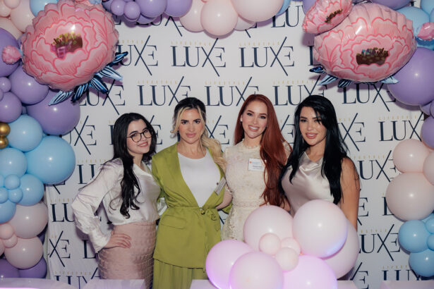 Four women stand smiling in front of a "LUXE" step-and-repeat banner, surrounded by pastel balloons and large pink floral balloon decorations at an event. South Florida Business & Wealth