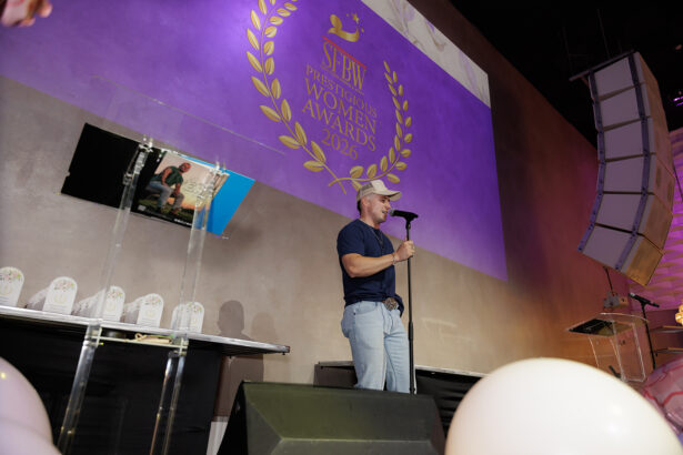 A man in jeans and a cap speaks into a microphone on stage at the SFBW Prestigious Women Awards 2026. The event logo is projected behind him, and awards are displayed on a table to his left. South Florida Business & Wealth