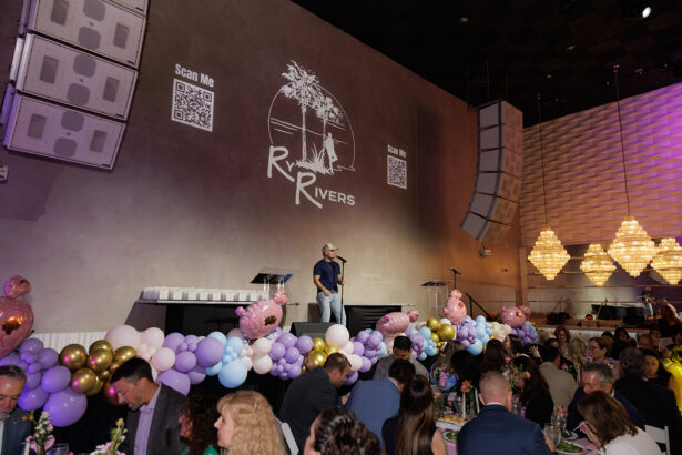 A man speaks or sings on a decorated stage at an indoor event with people seated at tables. The wall behind him displays a logo reading "Rivers" with palm trees, QR codes, and festive balloons along the stage. South Florida Business & Wealth