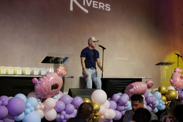 A man in a cap, t-shirt, and jeans stands at a microphone on stage decorated with pastel balloons and floral balloons, with two podiums and an audience in the foreground. South Florida Business & Wealth