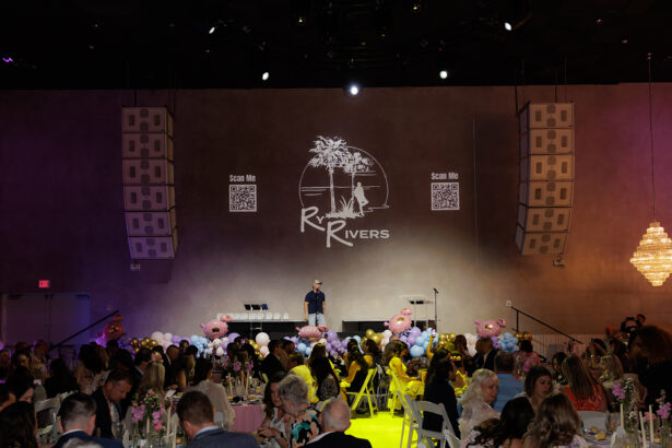A formal indoor event with seated guests at decorated tables. A person stands on stage under a projected "Rivers" logo, QR codes, and palm tree graphic. Balloon decorations and a chandelier are visible. South Florida Business & Wealth