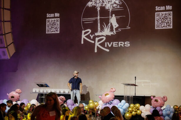 A man in a cap stands on stage in front of a wall with a large "Ry Rivers" logo, QR codes, and "Scan Me" text. The stage is decorated with pink flamingos and colorful balloons; people are seated at tables in the foreground. South Florida Business & Wealth