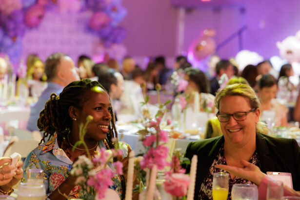 Two women smile and talk at a decorated event table with flowers and drinks, surrounded by other guests in a softly lit, festive setting. South Florida Business & Wealth