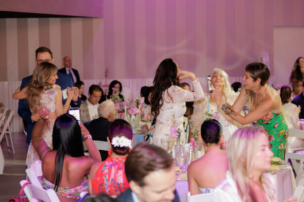 A lively indoor celebration with guests in colorful attire, some standing and taking photos, others sitting at decorated tables with flowers, while a few people embrace and smile in the foreground. South Florida Business & Wealth