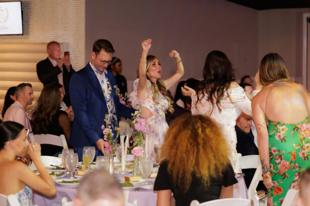 A woman stands with her arms raised in excitement while others sit or stand around a banquet table at a formal event. People are dressed in semi-formal attire, and there are flowers and drinks on the table. South Florida Business & Wealth