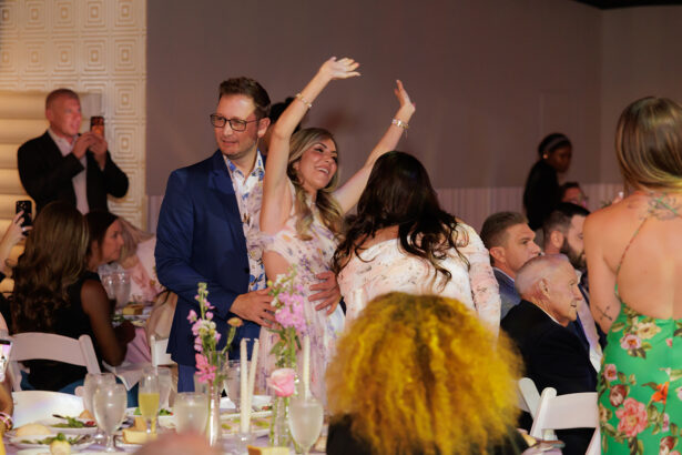 A woman in a floral dress joyfully raises her arms while standing next to a man in a blue suit at a crowded, festive indoor event with seated guests and decorated tables. South Florida Business & Wealth