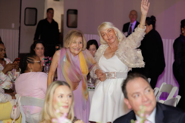 Two older women smile while standing among seated guests at a formal event. One woman waves and wears a white dress with a sparkly jacket; the other wears a pastel shawl. Other guests are seated around decorated tables. South Florida Business & Wealth