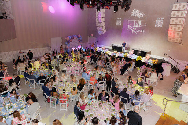 A large group of people sit at decorated round tables in a modern event hall with pastel balloons, a DJ booth, and projected logos and QR codes on the wall. The event has a festive, elegant atmosphere. South Florida Business & Wealth