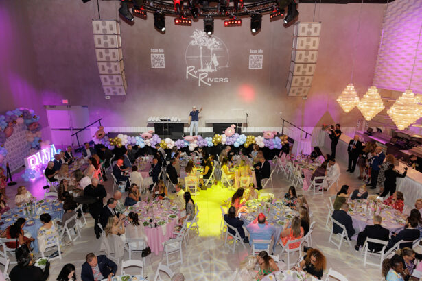 A large event in a modern, decorated hall with round tables, seated guests, and a stage. Balloons and pastel colors adorn the front. A speaker stands on stage; chandeliers and light projections add to the festive atmosphere. South Florida Business & Wealth