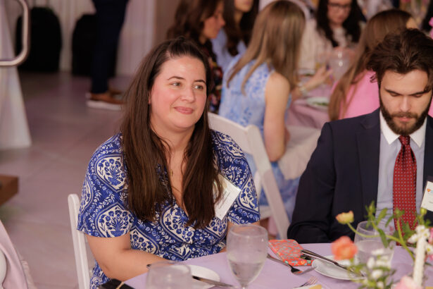 A woman in a blue patterned shirt sits at a round table with other people at a formal event. She is smiling slightly, and there are glasses, plates, and flowers on the table. South Florida Business & Wealth