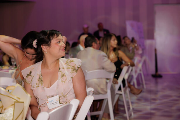 A woman in a light floral dress sits at a table, smiling and looking to the side, during a formal indoor event. Other attendees are seated around her, and the room is softly lit with purple tones. South Florida Business & Wealth