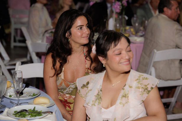 Two women sitting at a table during a formal event, smiling and looking toward the front. The table is set with plates of food, glasses, and bread rolls. Other guests are visible in the background. South Florida Business & Wealth