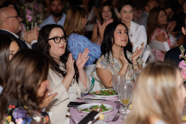 A group of people seated at a table with plates of salad, clapping and smiling during an indoor event. The atmosphere appears festive, with floral decorations on the table. South Florida Business & Wealth