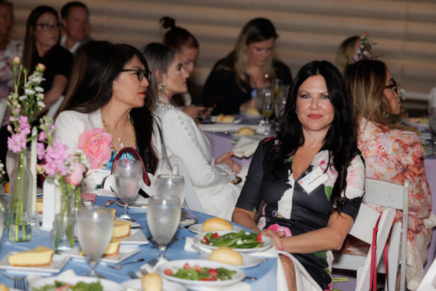 Several women sit at decorated tables with plates of salad, bread, and glasses of water, attentively listening at what appears to be a formal event or banquet. Floral centerpieces and pastel colors decorate the setting. South Florida Business & Wealth