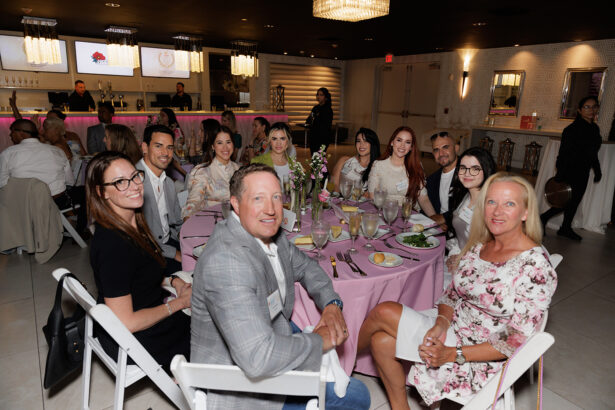 A group of people dressed in semi-formal attire sit around a table with pink tablecloth at an indoor event, smiling at the camera. Plates, glasses, and floral centerpieces are on the table. South Florida Business & Wealth
