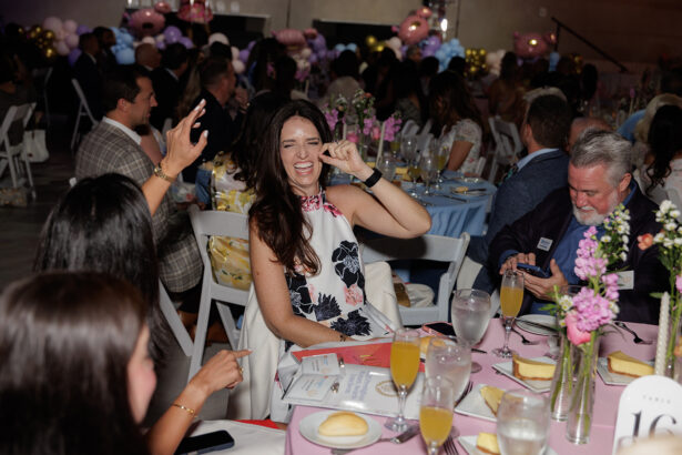 A woman in a white floral dress laughs and gestures at a table set for a formal event, surrounded by people, flowers, and drinks. The atmosphere is lively, with party decorations and guests in the background. South Florida Business & Wealth