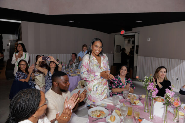 A woman in a floral dress stands and smiles as people seated around her clap; others take photos. The group sits at a decorated table with flowers, plates of food, and drinks in a softly lit event space. South Florida Business & Wealth
