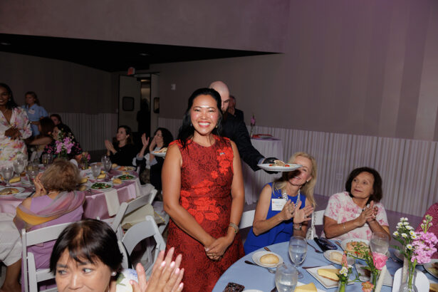 A woman in a red dress smiles while standing in a banquet hall, surrounded by seated guests who are clapping. The atmosphere is cheerful, with several tables decorated for a celebratory event. South Florida Business & Wealth