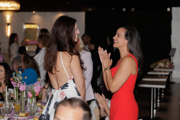 Two women at a lively indoor event are smiling and conversing; one wears a red dress and claps her hands, while the other wears a backless white floral dress. People and tables with drinks and flowers are visible in the background. South Florida Business & Wealth