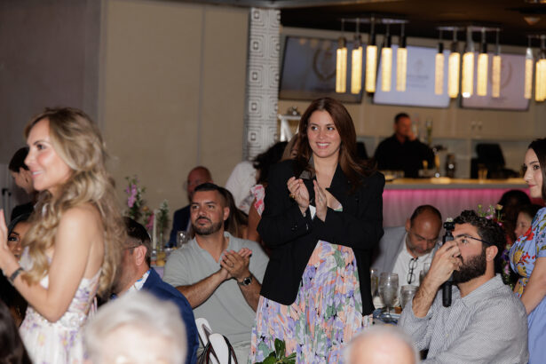 A woman stands and claps while others sit and watch at a formal indoor event. Some people are seated at tables with drinks and flowers, and soft lighting creates a warm atmosphere. South Florida Business & Wealth